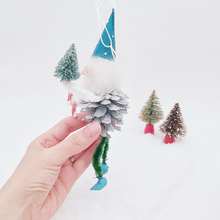 Load image into Gallery viewer, Opposite side view of spun cotton pine cone elf, held in hand against a white background with two vintage bottle brush trees in distance. Pic 6 of 7. 
