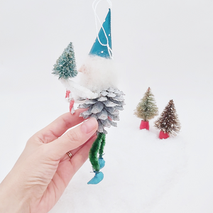 Opposite side view of spun cotton pine cone elf, held in hand against a white background with two vintage bottle brush trees in distance. Pic 6 of 7. 