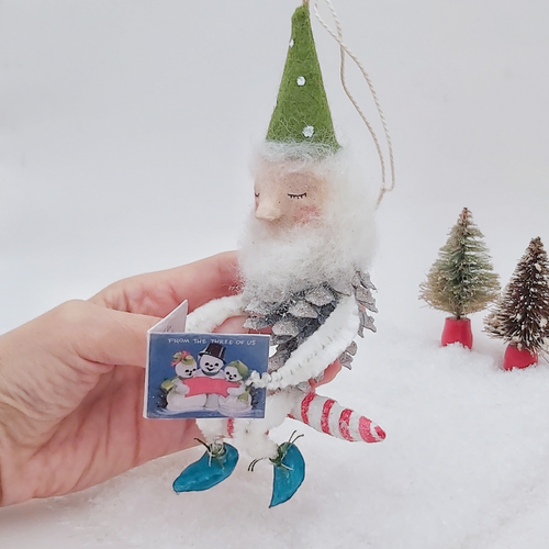 Vintage style spun cotton pine cone elf sitting on a spun cotton candy cane. He's held in hand against a white background with vintage bottle brush trees in the distance. Pic 1 of 10. 