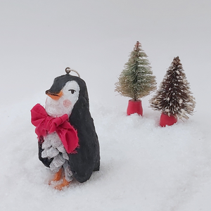 Vintage style spun cotton pine cone penguin ornament, standing on fake snow against a white background. Two miniature bottle brush trees are in the distance. Pic 5 of 10.
