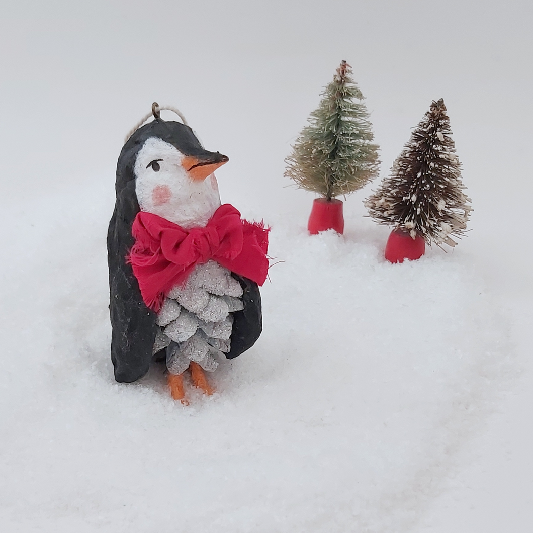 A front view of spun cotton pine cone penguin standing on fake snow with two miniature bottle brush trees in the distance. Pic 1 of 10.