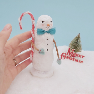 Image of a hand next to a vintage style spun cotton skinny snowman. He stands on snow against a light blue background. Pic 2 of 6. 