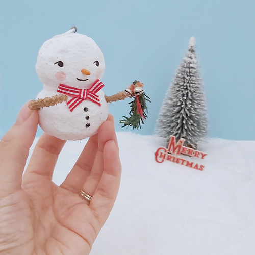 Vintage style spun cotton snowman ornament, held in hand for size comparison. He's against a light blue background with a bottle brush tree and vintage Merry Christmas sign in the distance. Pic 1 of 7.