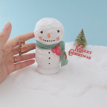 Cargar imagen en el visor de la galería, A hand next to the vintage style spun cotton snowman, for a size comparison. He sits on snow against a light blue background, in front of a vintage bottle brush tree and Merry Christmas sign. Pic 2 of 7.
