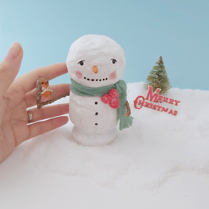 A hand next to the vintage style spun cotton snowman, for a size comparison. He sits on snow against a light blue background, in front of a vintage bottle brush tree and Merry Christmas sign. Pic 2 of 7.