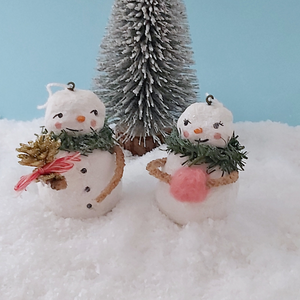 Vintage style spun cotton snowman and snow lady ornament set, sitting on white snow against a light blue background. A bottle brush tree sits behind them. Pic 1 of 11. 