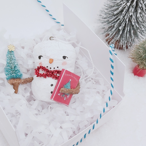 Vintage style spun cotton snowman ornament laying in white gift box with white shredding, next to bottle brush trees on a white background. Pic  5 of 8.