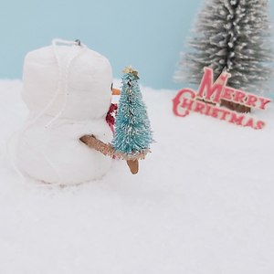 Side view of vintage style spun cotton snowman, sitting on snow with bottle brush tree and Merry Christmas sign  in distance. Pic 7 of 8.