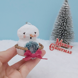 Vintage style spun cotton snowman ornament, held in hand against a light blue background with a bottle brush tree and Merry Christmas decoration in the distance. Pic  2 of 8.
