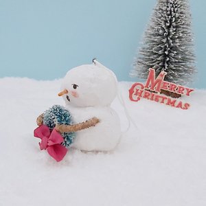 Opposite side view of vintage style spun cotton snowman. He's against a light blue background, sitting on snow, with a bottle brush tree in the distance. Pic  7 of 8.